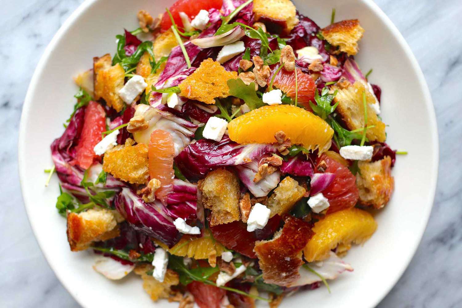 Horizontal overhead view of citrus salad on a white plate. Supremed orange and grapefruit, radicchio, croutons and feta cheese crumbles are visible.