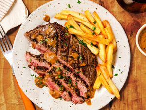 Overhead view of a platter of partially sliced flank steak and french fries next to a small bowl of sauce and fork all on a wooden tabletop
