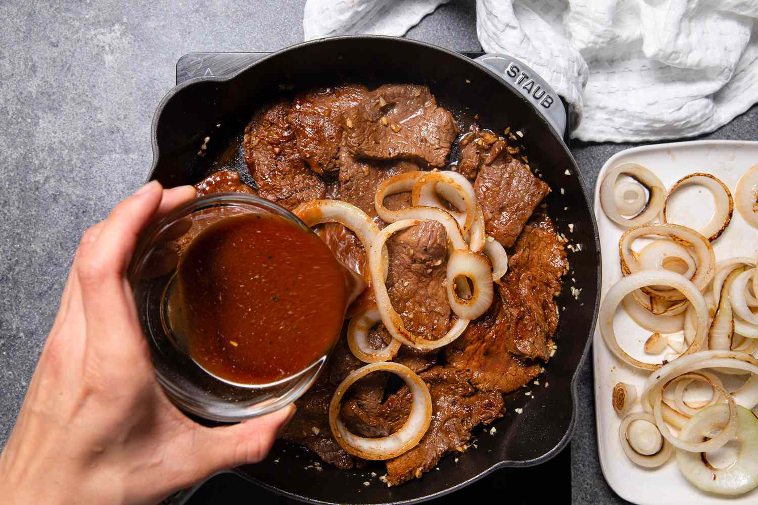 Marinade Added to the Bistek and Onions in the Cast Iron Skillet, and Next to It, a Plate With Cooked Onions and a White Kitchen Towel 