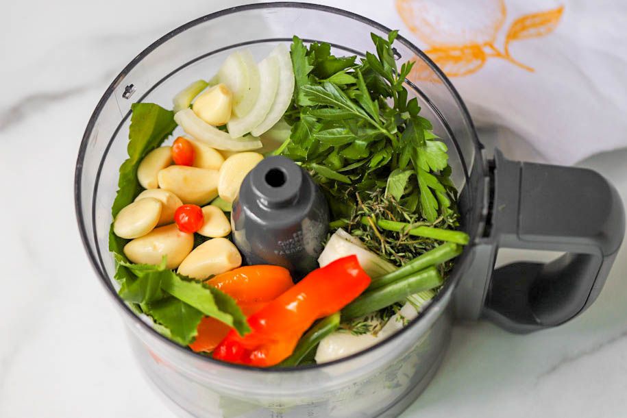 In a food processor: peppers, culantro, thyme, green onions, halved white onion, garlic, basil, and parsely for Carribbean green seasoning recipe