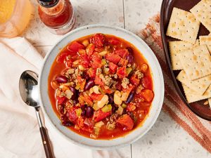 Bowl of copycat Wendy's chili at a table setting with a bowl of saltines on a brown and white kitchen towel, a bottle of tabasco, a drink, and a white table napkin