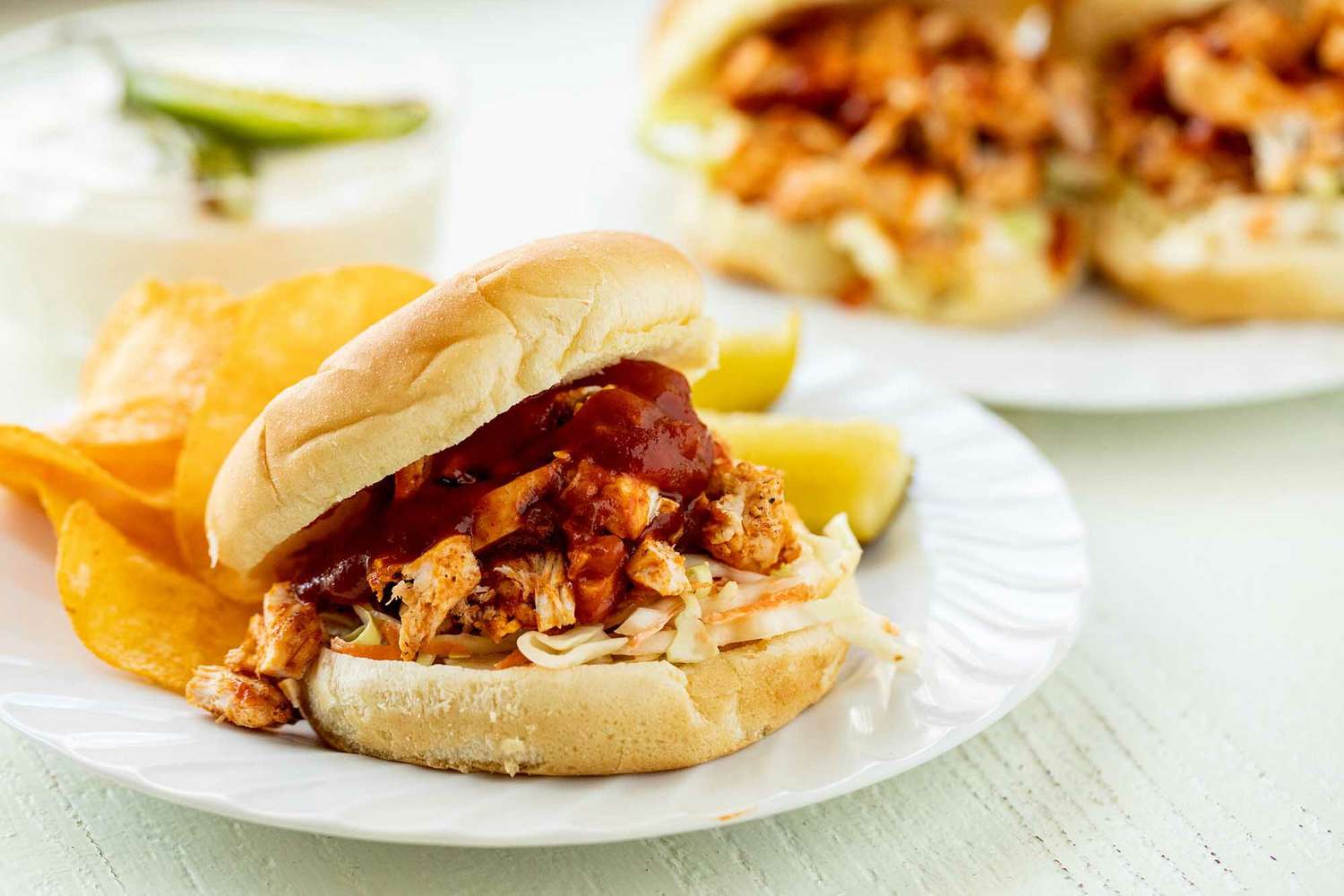 A plate with pickles, chips and shredded bbq chicken sandwich with pickle slaw and topped with bbq sauce. A second plate with two sandwiches and sarrano mayo are behind the plate.
