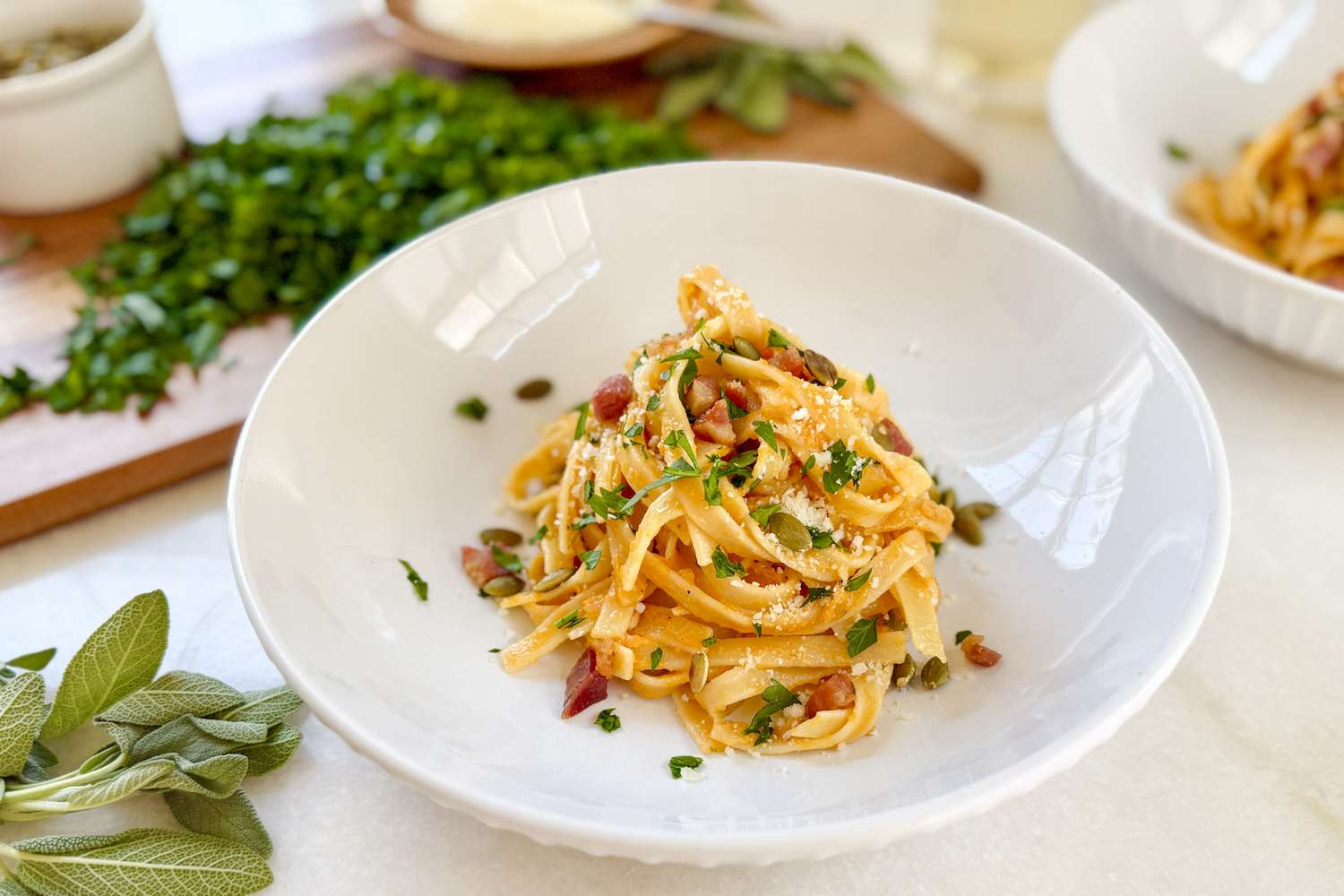 A plate of pasta garnished with herbs and small bits of an ingredient on a white surface with other meal components in the background