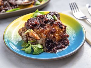 A plated chicken dish topped with a sticky sauce and garnished with herbs with a fork and knife visible in the background