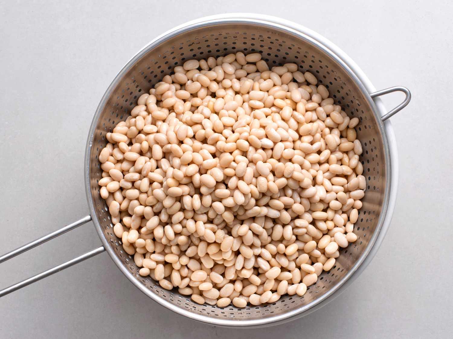 Soaked and drained white beans in a metal colander on a white stone background