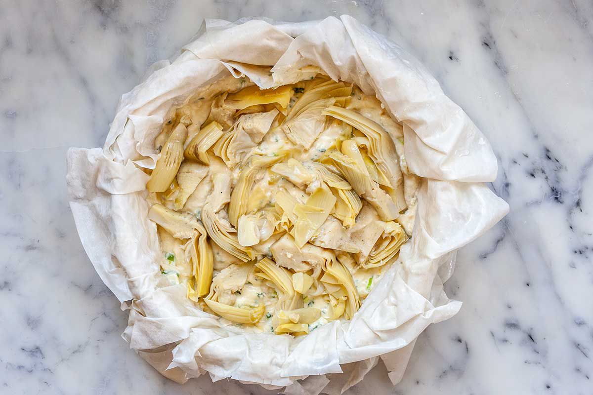 A cake pan is on a marble countertop. Unbaked phyllo dough is layered underneath a layer of chopped, canned artichokes. The phyllo is draped over the sides of the pan.