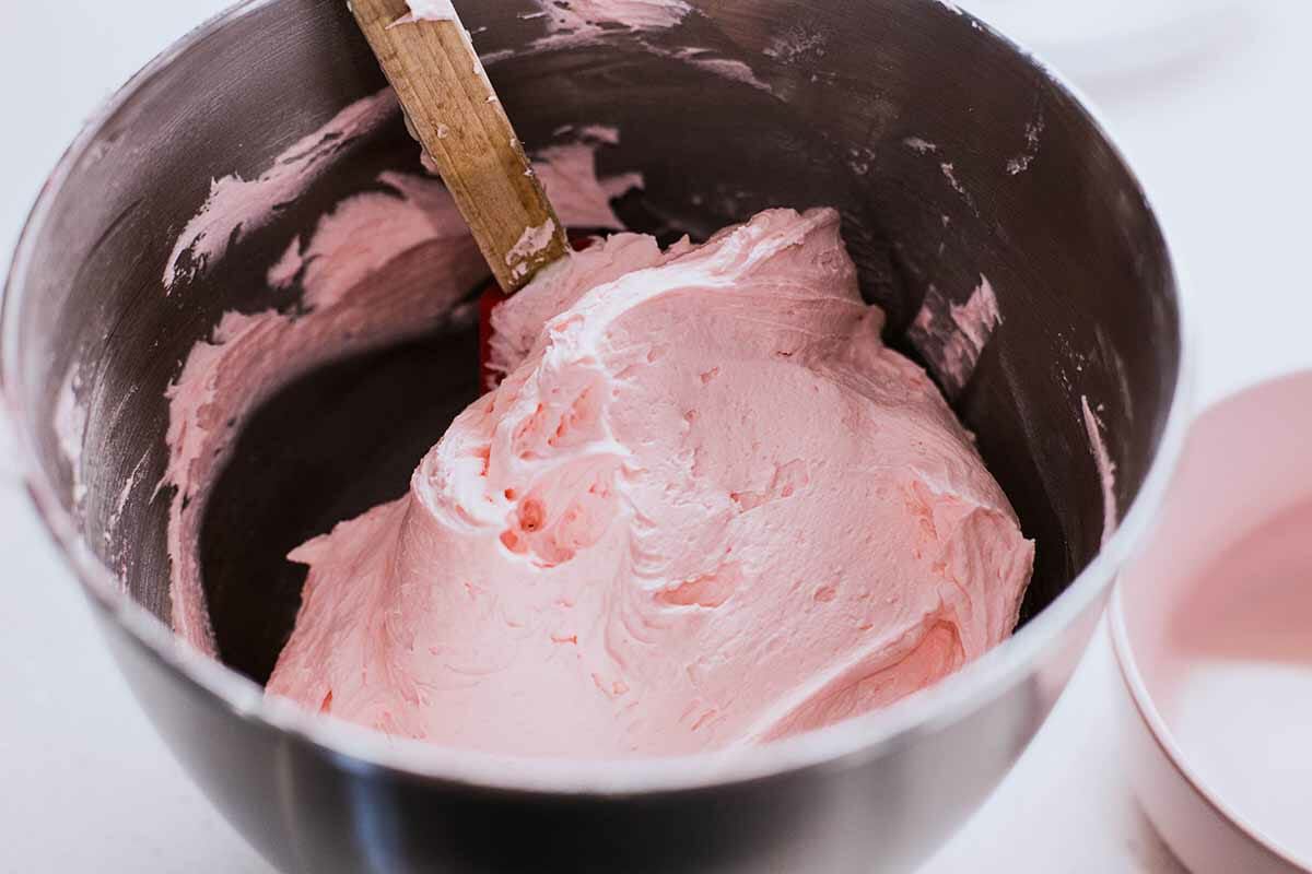 A big stand mixer bowl of pink frosting with a spatula in it