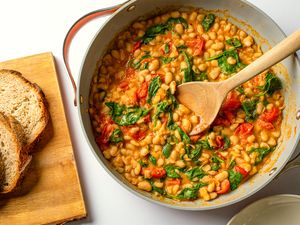 Pan of stewed beans with spinach and tomatoes wooden spoon and sliced bread on a cutting board