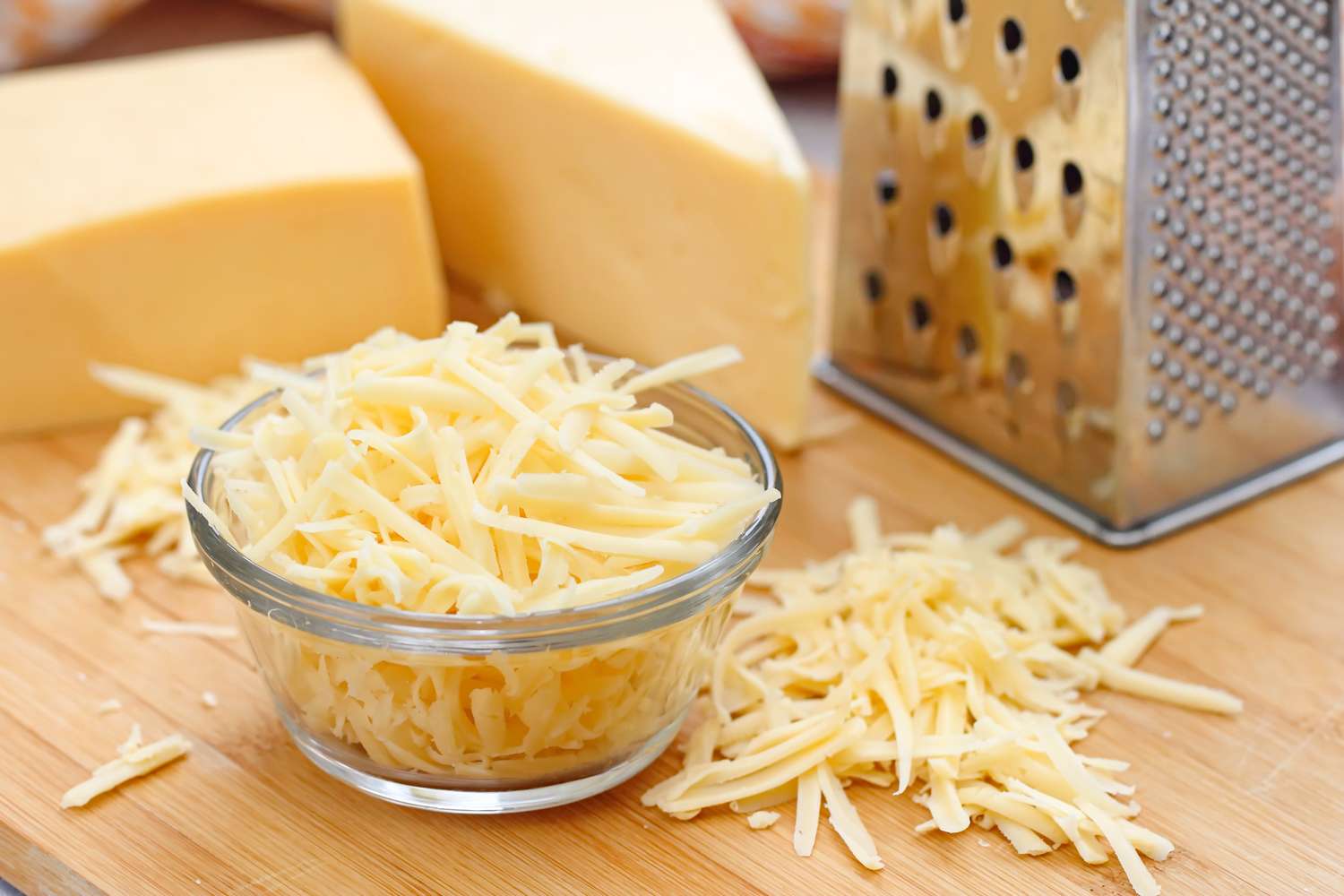 Grated cheese in a small bowl with cheese blocks and a grater in the background