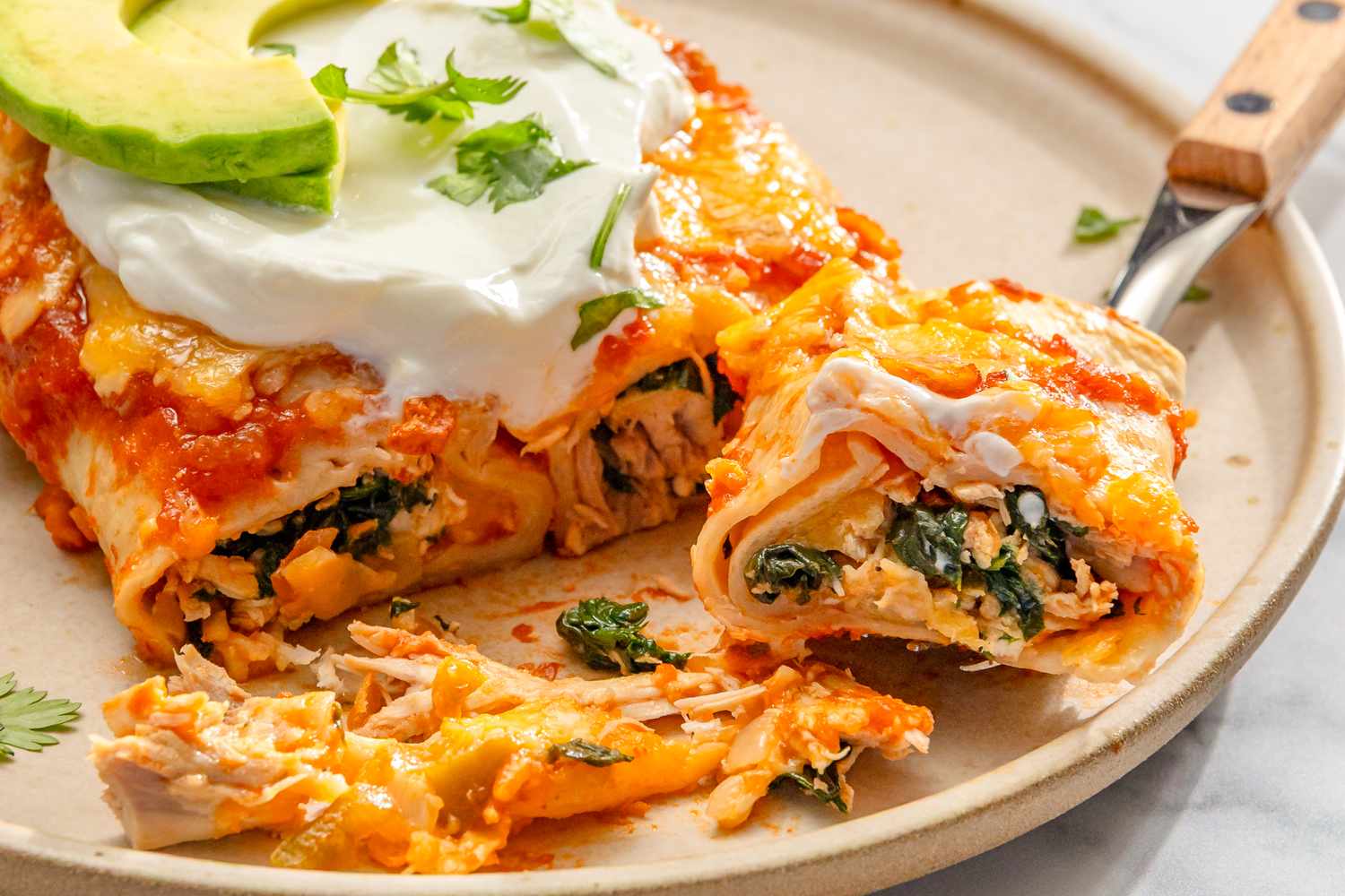 Chicken and spinach tortilla with melted cheese sliced avocado and sour cream on top a fork taking a bite on a plate