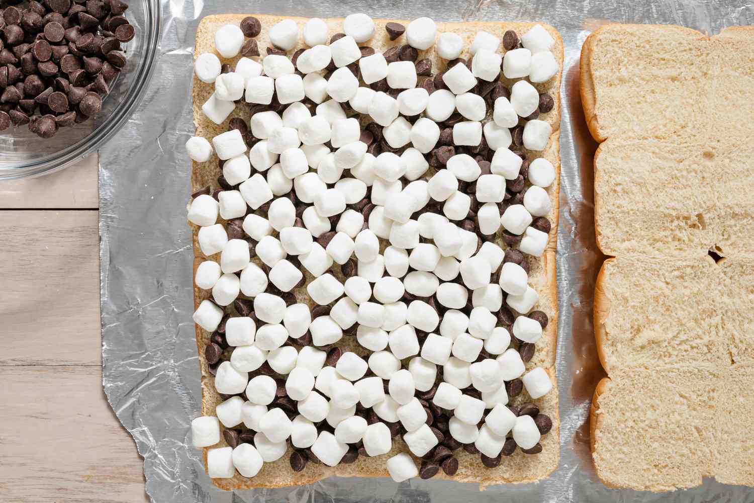 Chocolate Chips Topped With Mini Marshmallows on the Same Halved Slider Loaf. On the Left, a Bowl With More Chocolate Chips and on the Right, the Other Halved Slider Loaf for Grilled S'mores Slider Recipe