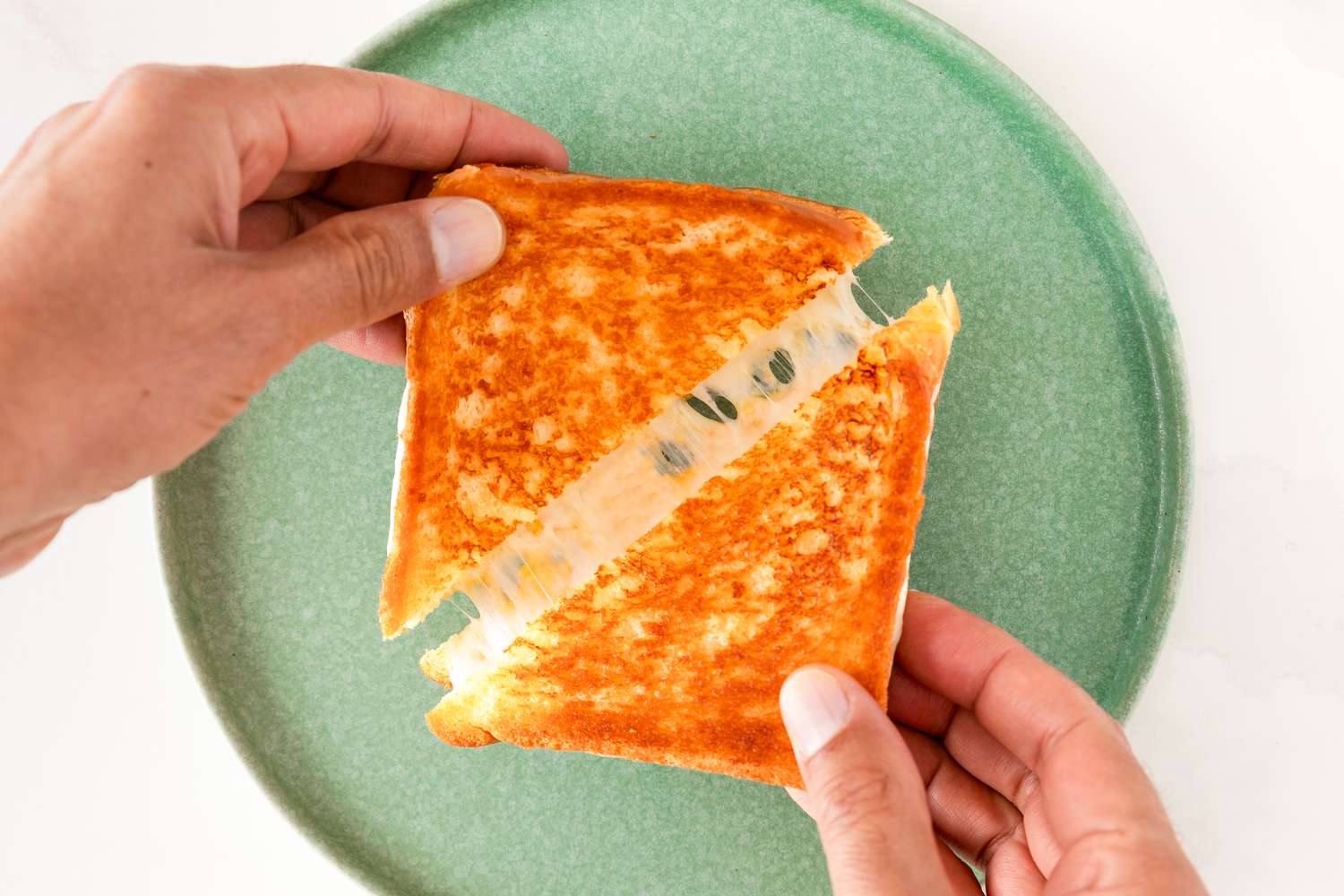 Hands pulling apart a grilled cheese sandwich on a green plate