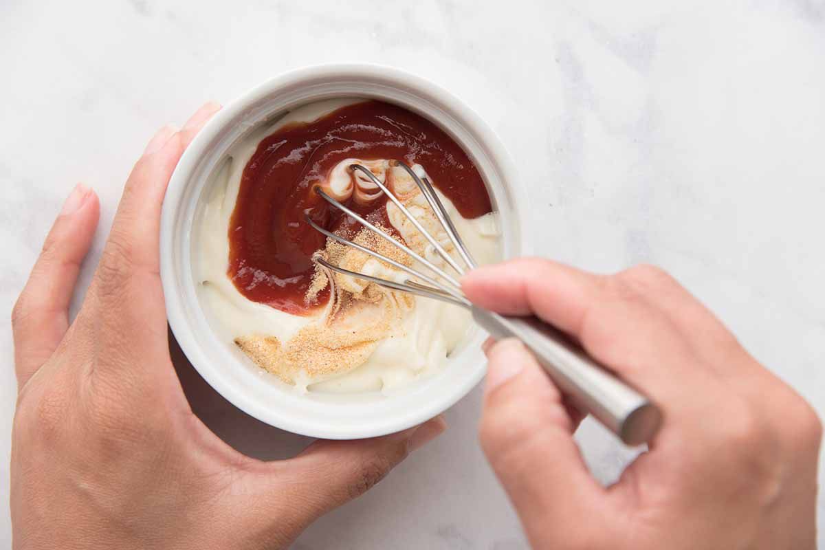 Person combining ketchup and mayo in a small bowl with a whisk.