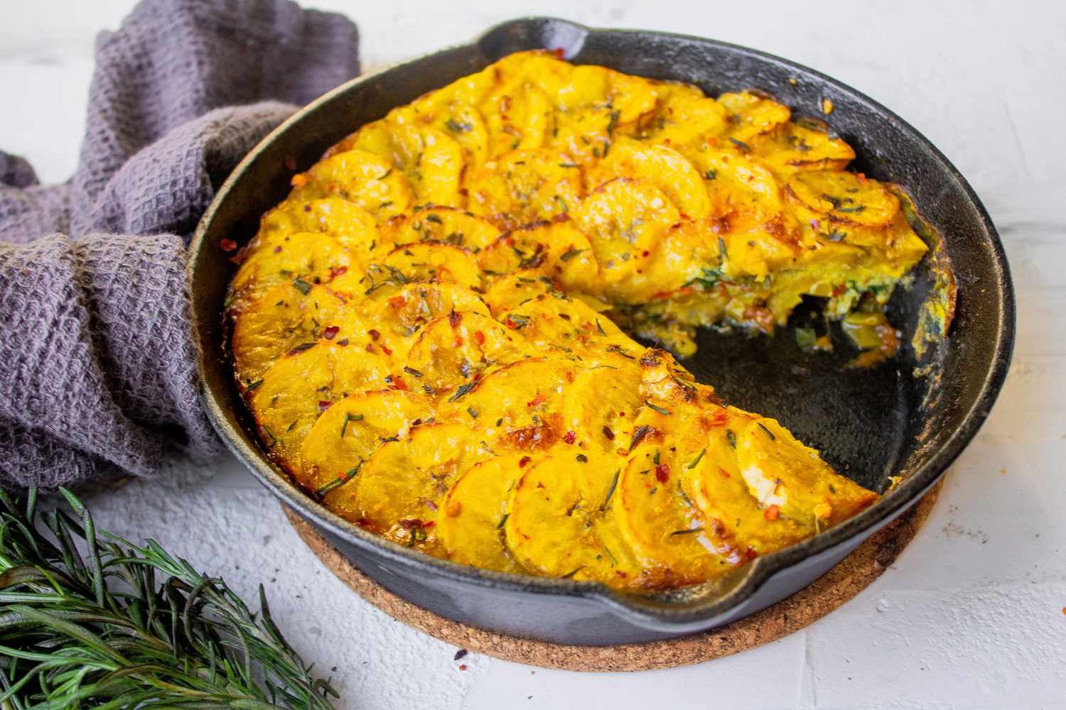 Plantain Frittata with a Serving Cut Out in Cast Iron Skillet Surrounded by Rosemary and Kitchen Towel