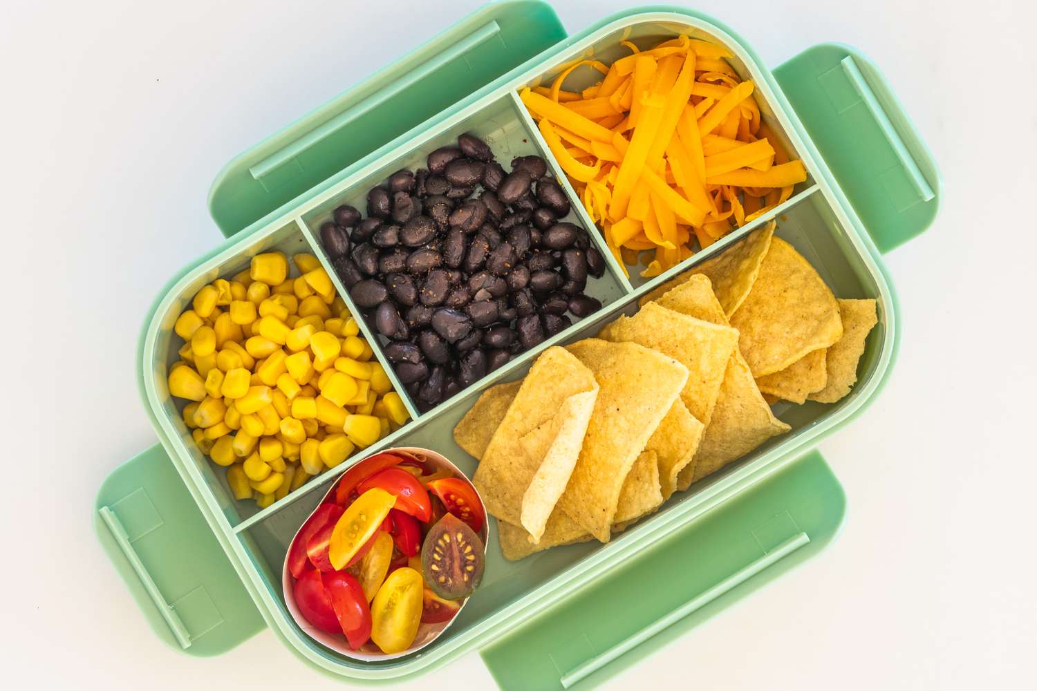 Overhead view of a bento-style lunch box with corn, black beans, shredded cheese, tomatoes and tortilla chips on a white background