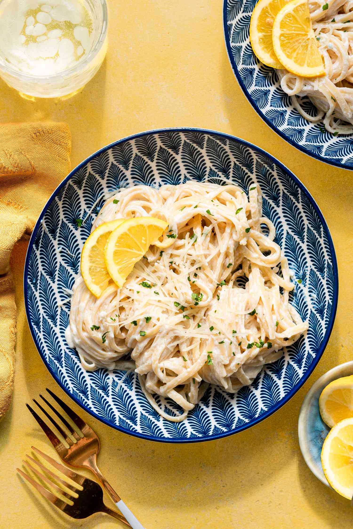 creamy lemon pasta in a bowl at a table setting with another serving in a plate, a glass of water, table napkin, and a bowl of sliced lemons, and forks