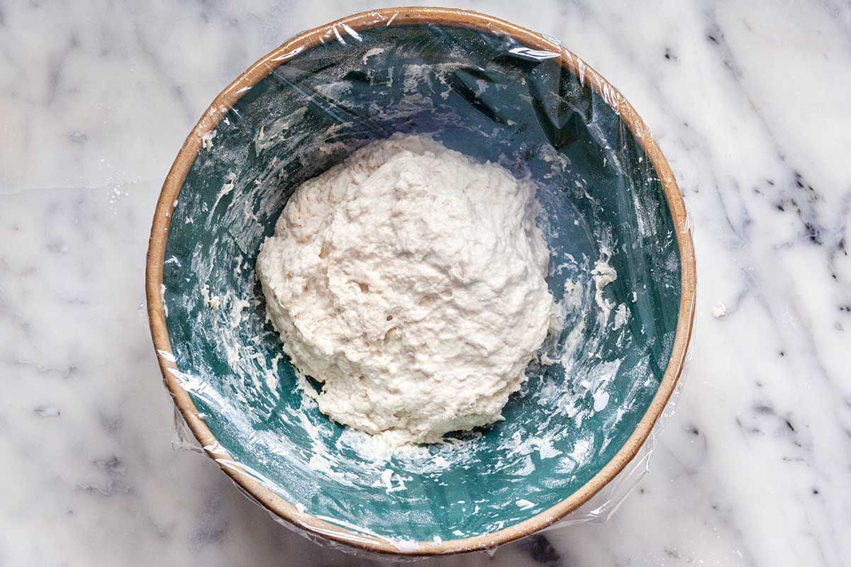 A bowl covered in plastic wrap with a wet dough inside to show how to make no knead bread.