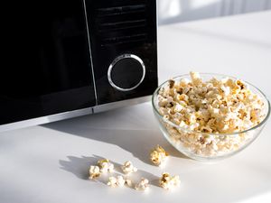 A bowl of popcorn on a counter next to a microwave some popcorn scattered outside the bowl