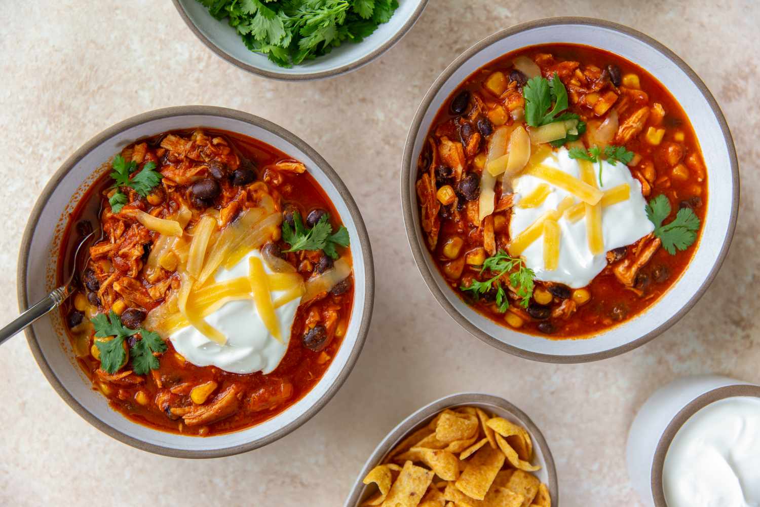 Bowls of chicken enchilada chili topped with sour cream and cilantro displayed with toppings and garnishes in surrounding bowls