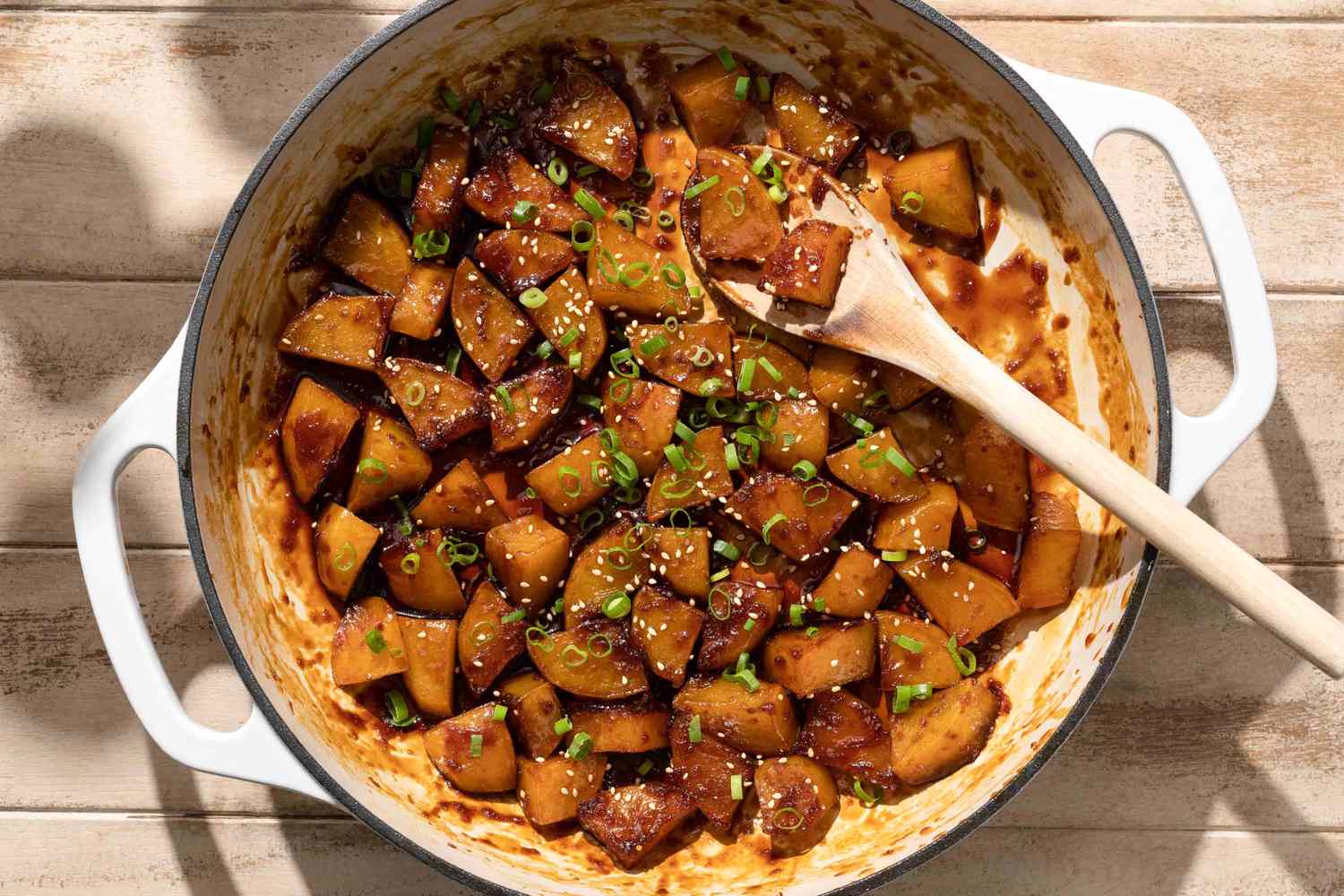 Braised glazed potatoes garnished with sliced green onions served in a pot with a wooden spoon