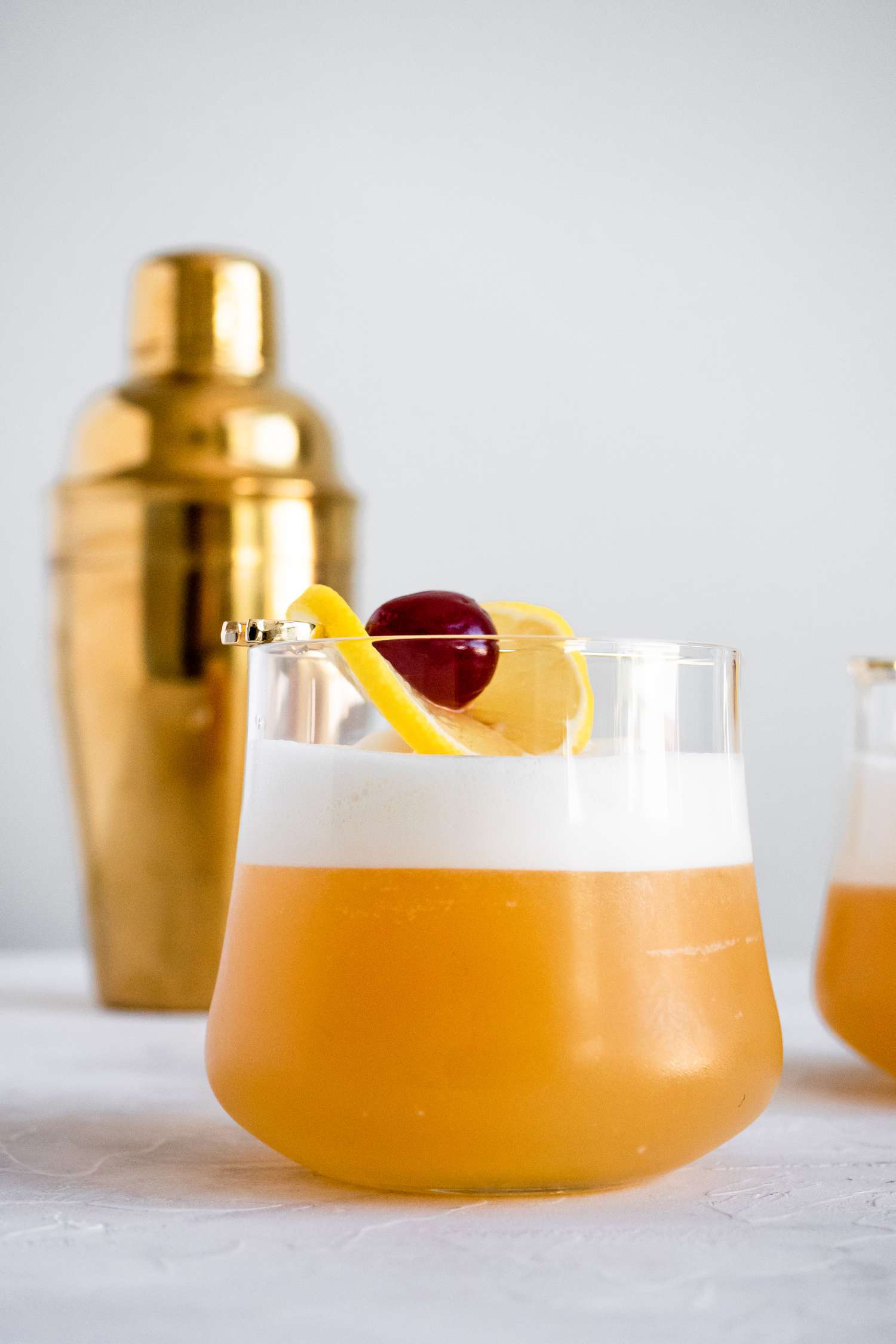 Amaretto Sour Garnished with Cherry and Lemon Slice with Shaker in Background