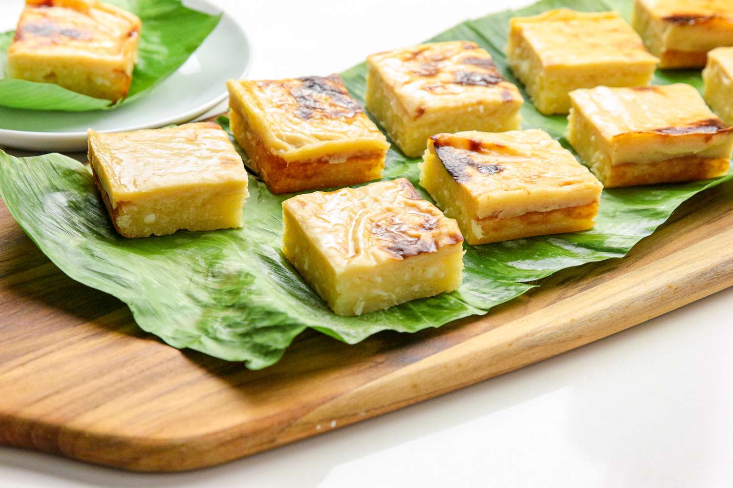 Cassava Cake Slices on a Banana Leaf Covered Wooden Tray, and in the Background, a Slice on a Banana Leaf Covered Plate