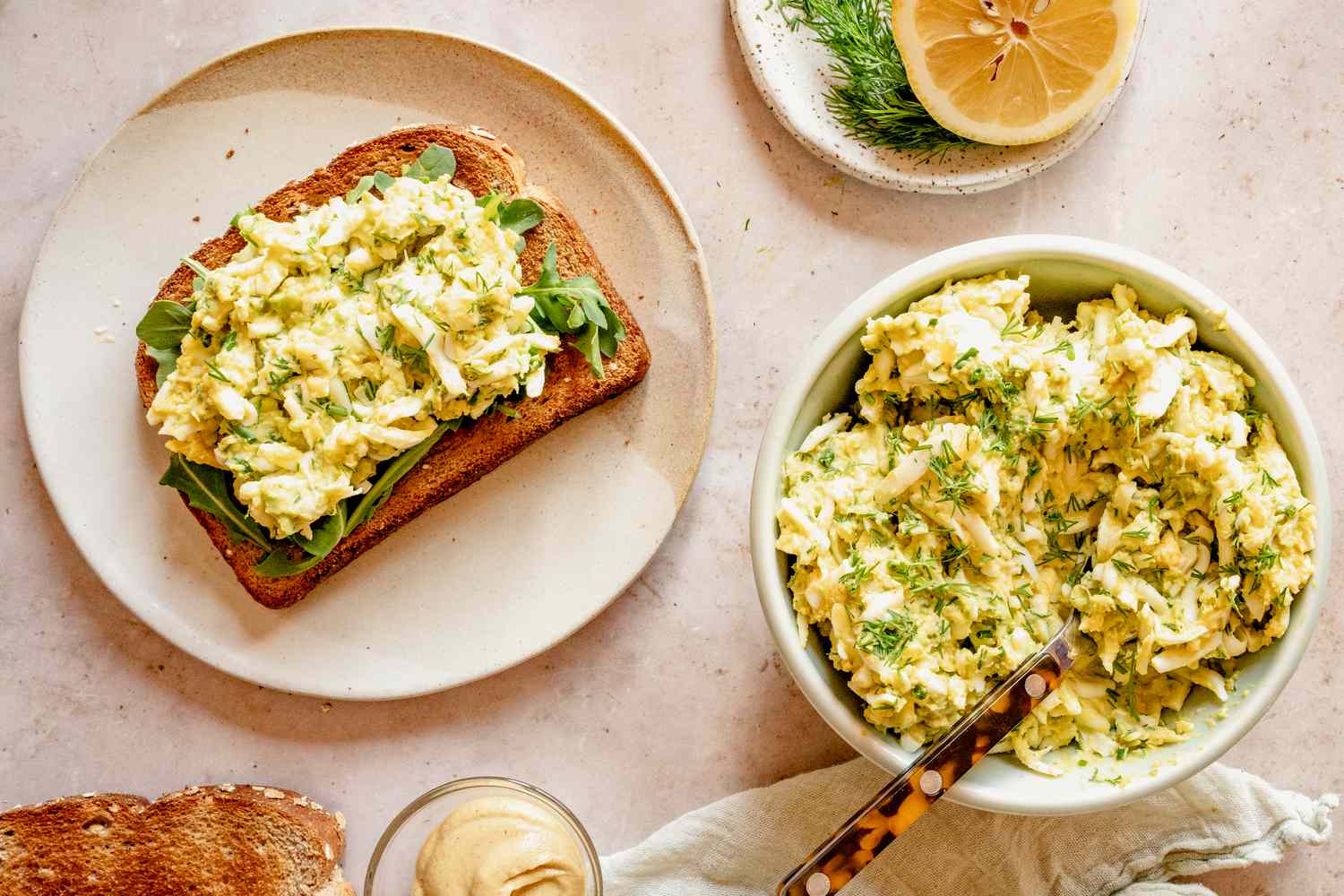 Overhead view of a bowl of mayo-free egg salad with a knife next to a small plate with a piece of toast covered in egg salad and greens