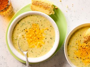 Overhead view of a two bowls of broccoli cheddar soup, one with a spoon and on a plate with pieces of bread, one with a piece of bread in the soup all next to a drink glass