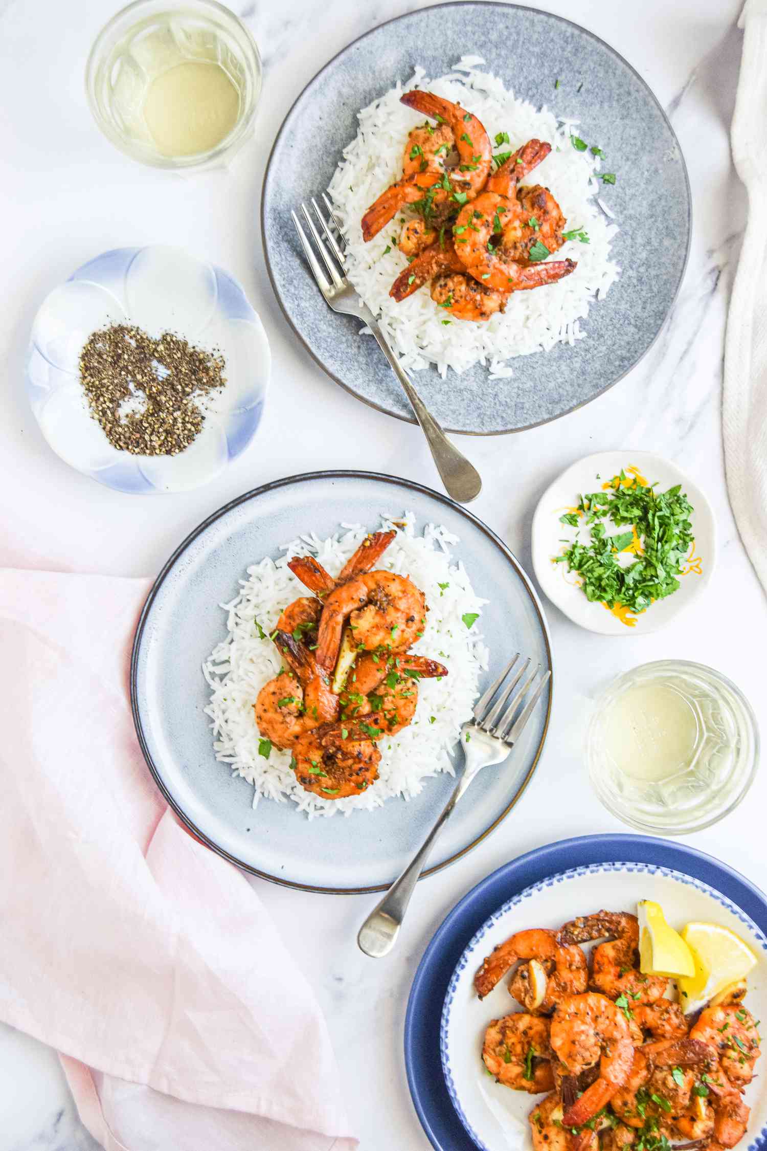 Two Plates of Peppery Garlic Shrimp Over Rice and Another Plate with Peppery Garlic Shrimp with Lemon Wedges, Surrounded by Glasses and Small Plates of Seasoning and Herbs 