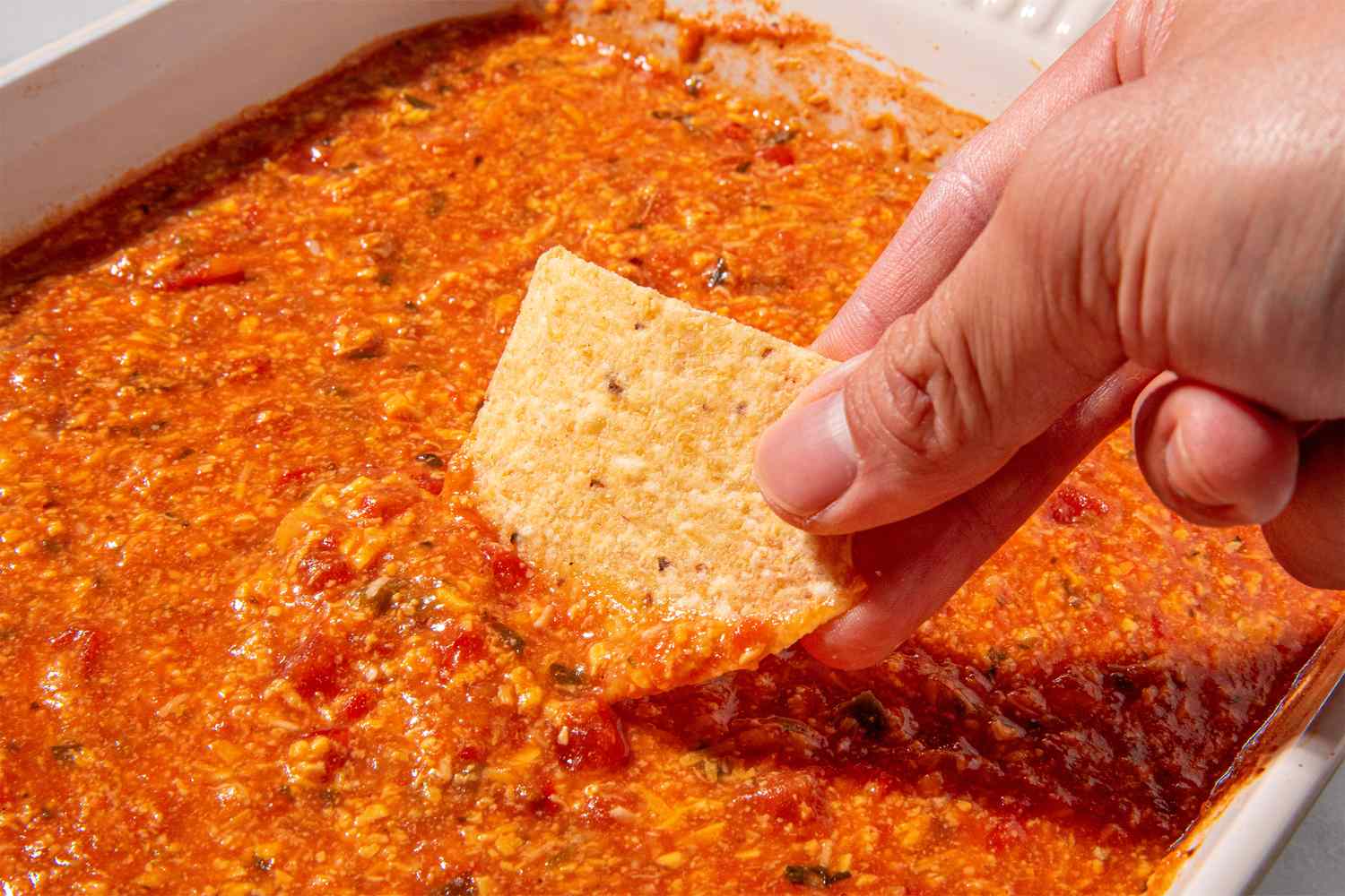 Hand dipping a tortilla chip into a tomato cheese dip in a dish