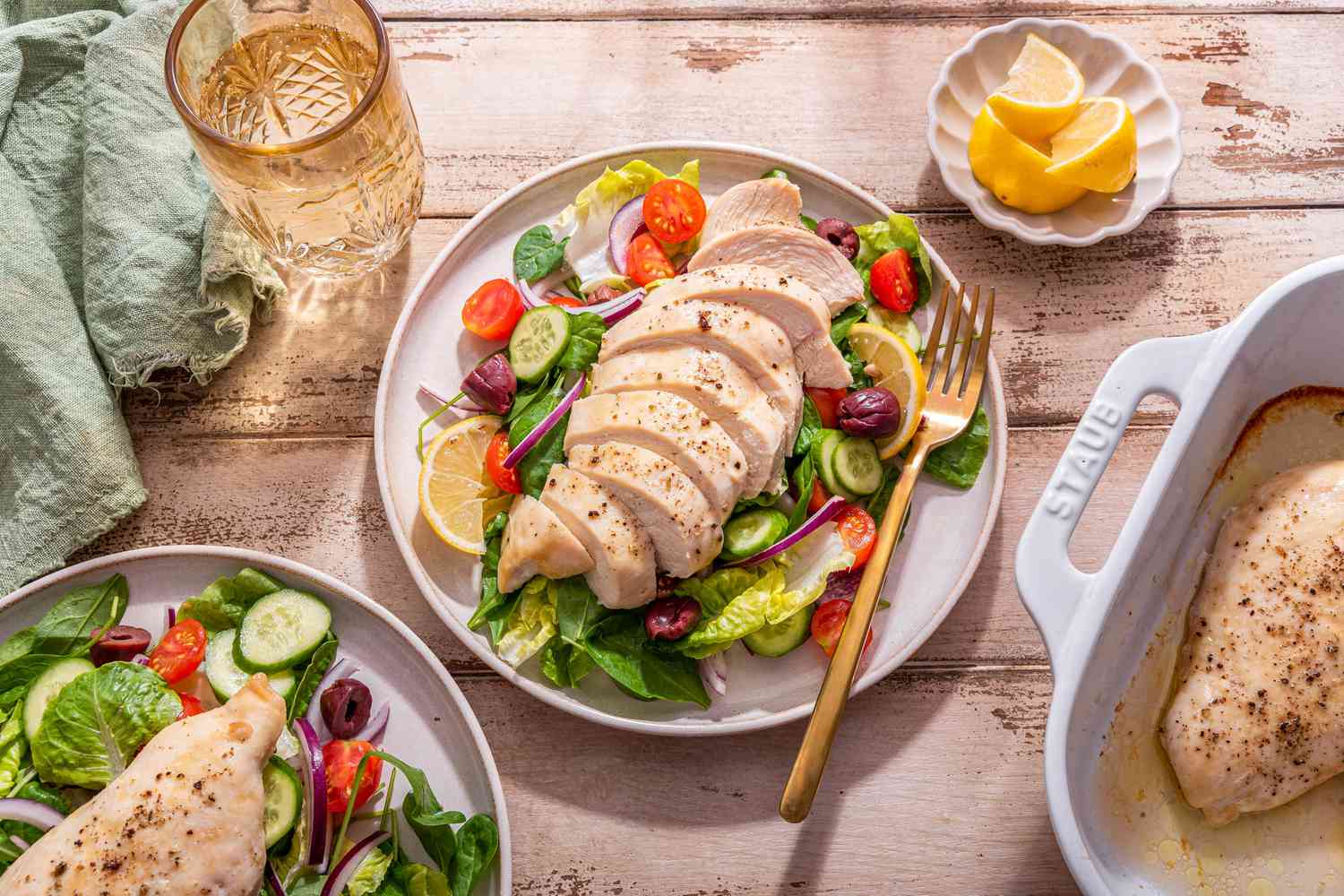 Sliced baked chicken breast on a plate with a salad, and in the surroundings, a casserole dish with more baked chicken, a small saucer with lemon slices, another serving of chicken and salad on a plate, a sage kitchen towel, and a glass of water