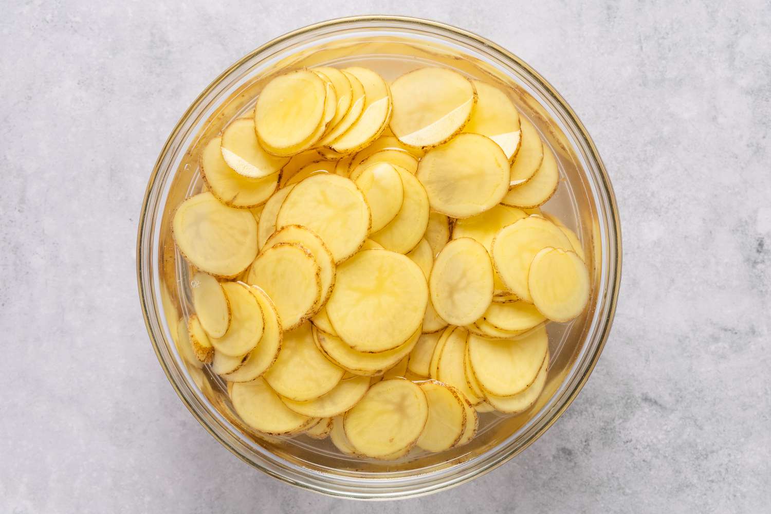 Overhead view of a clear glass bowl of potato slices in cold water for Sheet Pan Au Gratin Potatoes recipe