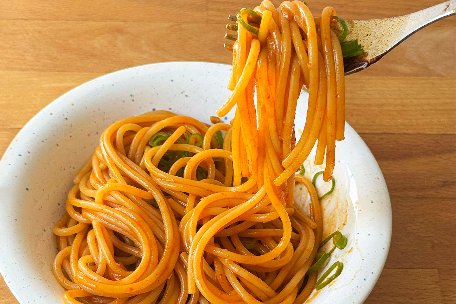 A bowl of cooked noodles being lifted by a fork garnished with green onion slices