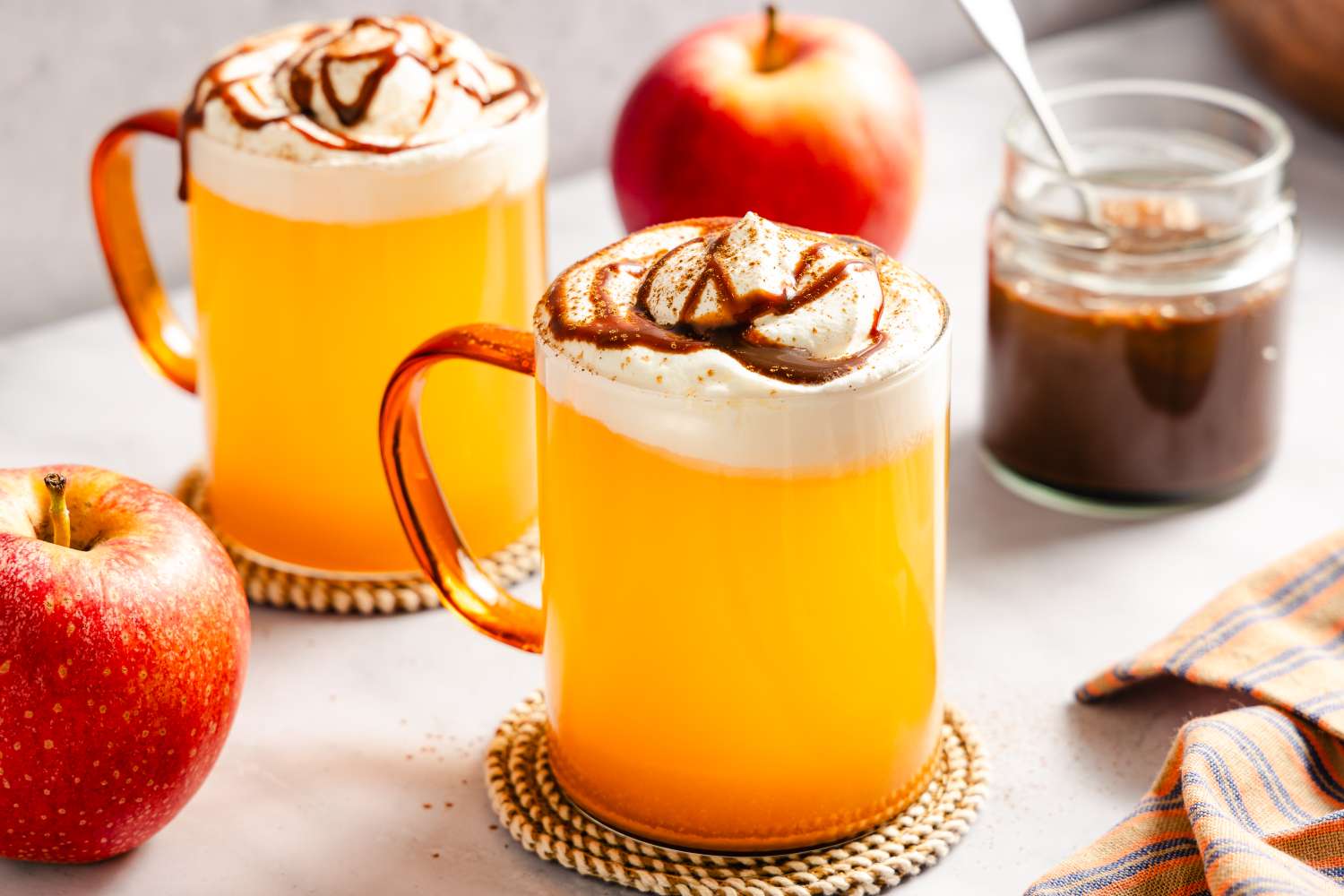 Two mugs of caramel apple cider topped with whipped cream accompanied by apples and a jar of caramel sauce