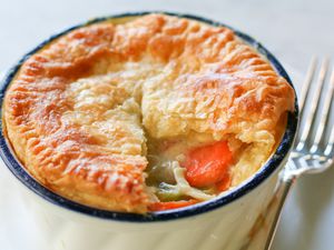 A pot pie with a golden crust is in a ramekin with some of the crust removed to show the filling.
