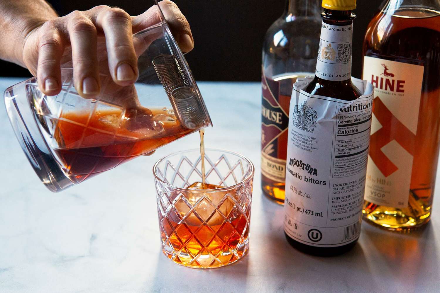A Vieux carre is being strained into a whiskey glass. Bottles for the cocktail are to the right.