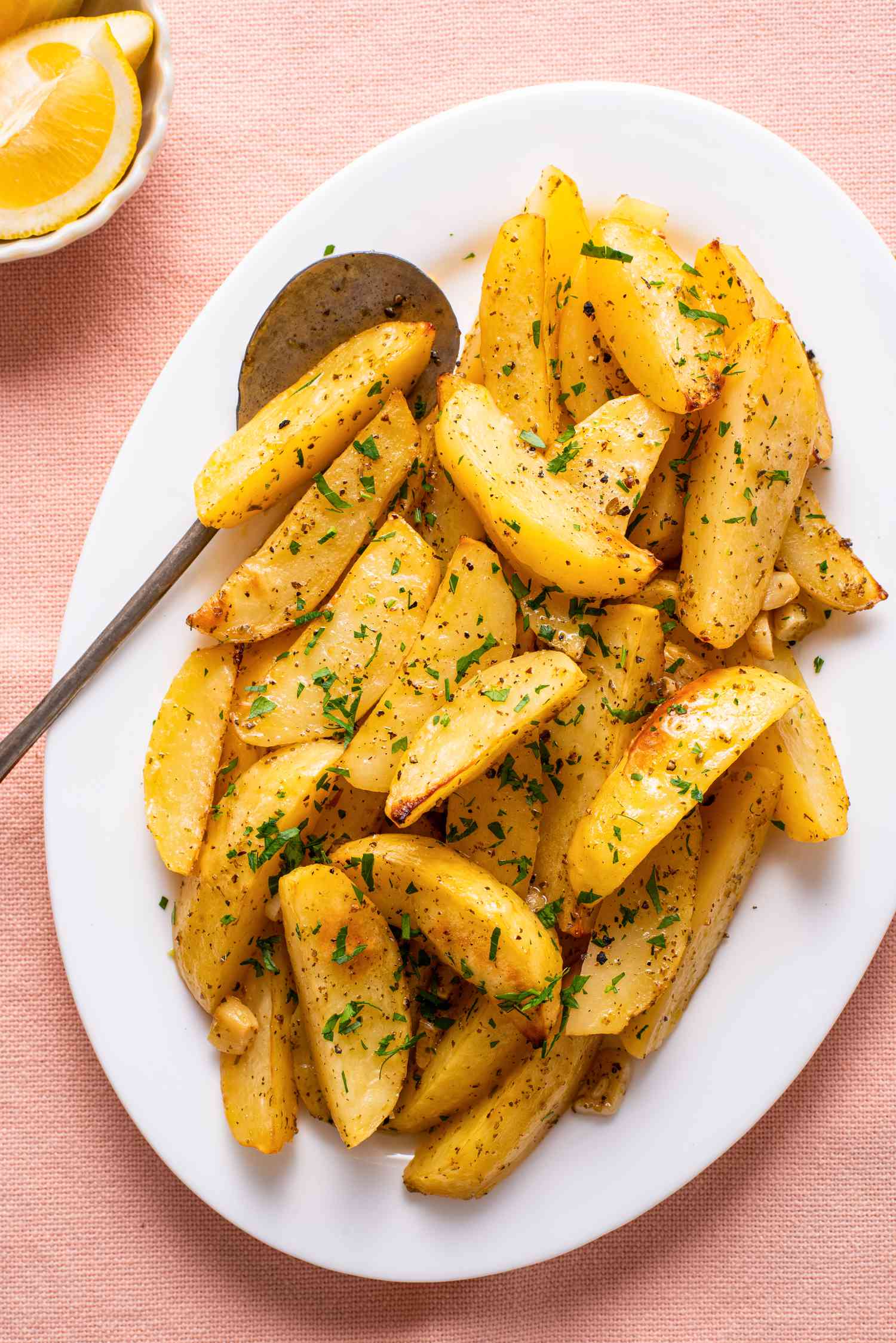greek lemon potatoes topped with minced herbs on a plate next to a small bowl of lemon wedges