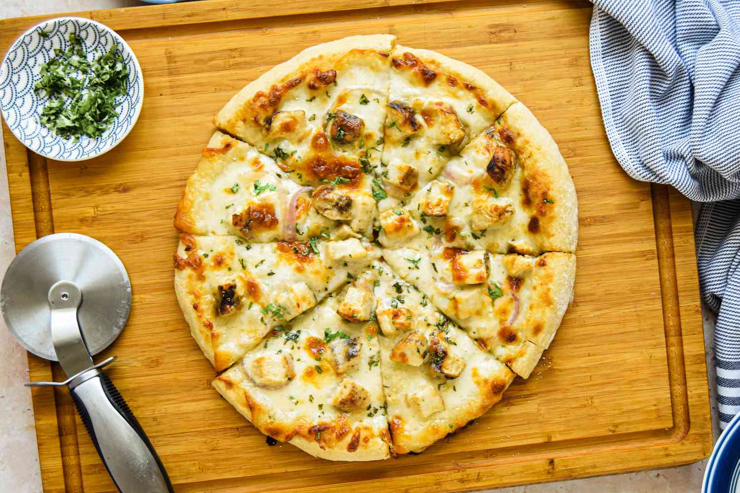 Chicken Alfredo Pizza Cut Into Slices Using a Pizza Roller on the Cutting Board, and in the Surroundings, a Small Bowl of Fresh Parsley and a Blue and White Striped Kitchen Towel