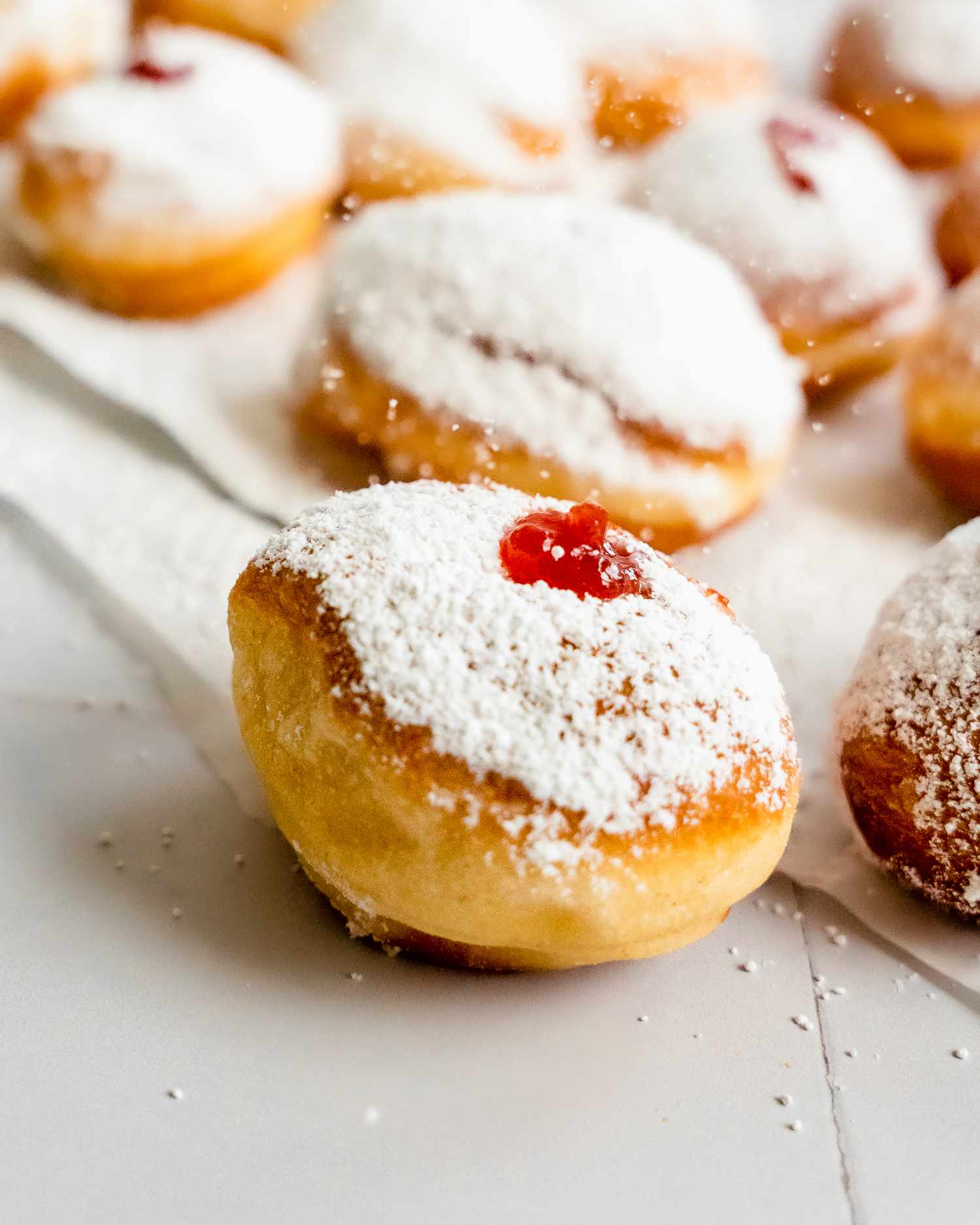 Powdered Sugar Sprinkled onto Sufganiyot (Israeli Jelly Donuts) on the Counter with More on the Parchment Paper Behind It 