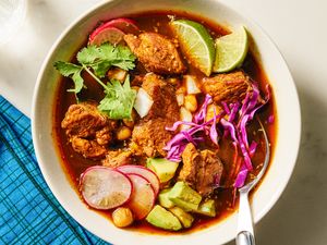 A bowl of pozole rojo garnished with lime wedges radish slices cabbage cilantro and pieces of pork placed on a blue tablecloth