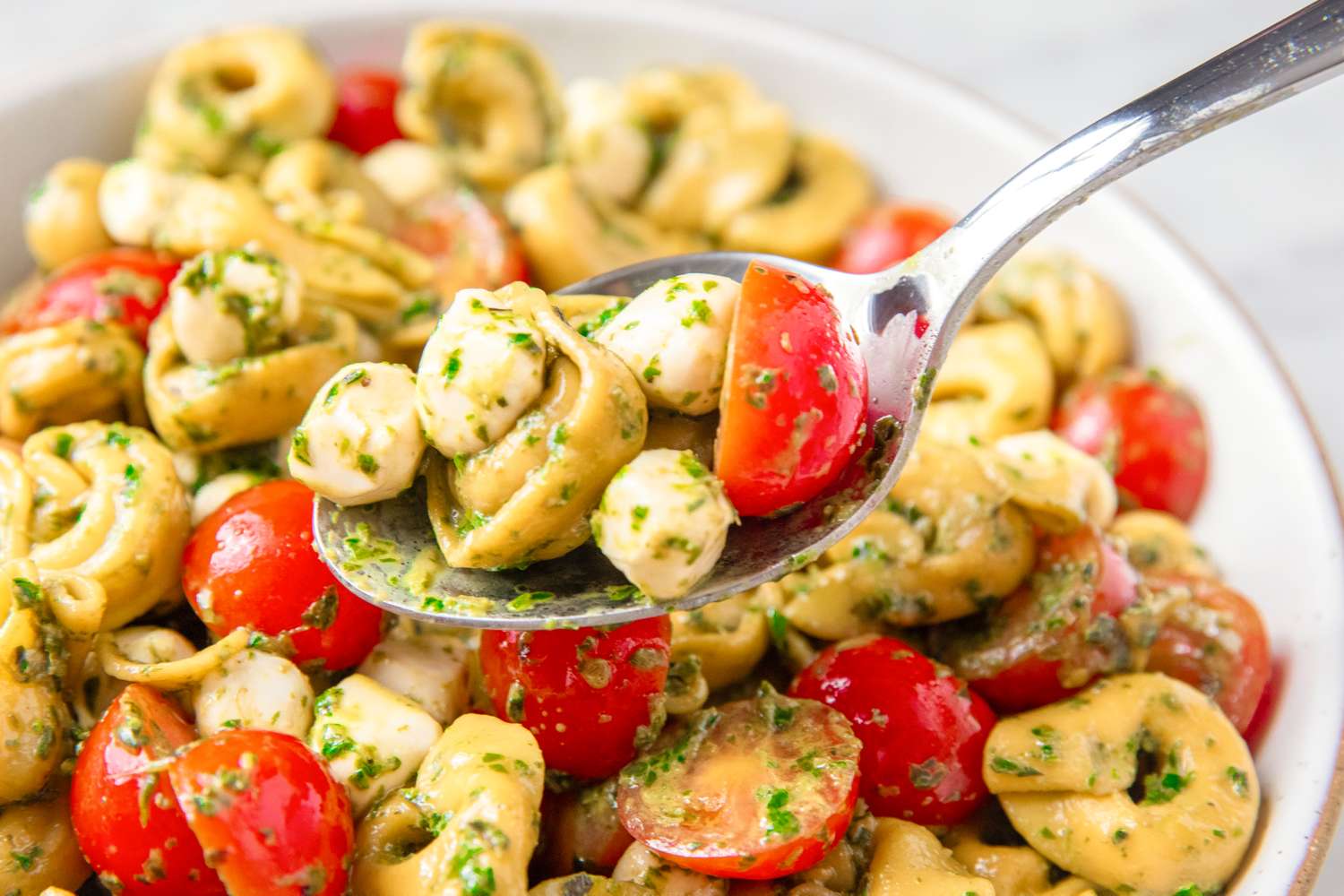 Tortellini salad with cherry tomatoes, mozzarella balls, and pesto on a plate, a spoon lifting a portion