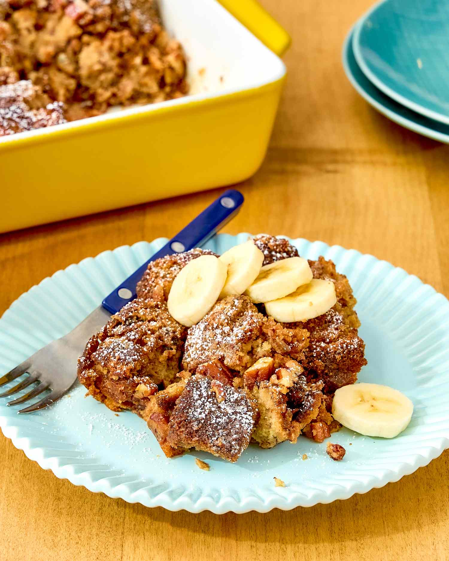 A serving of banana bread breakfast casserole topped with banana slices on a plate with the casserole dish in the background