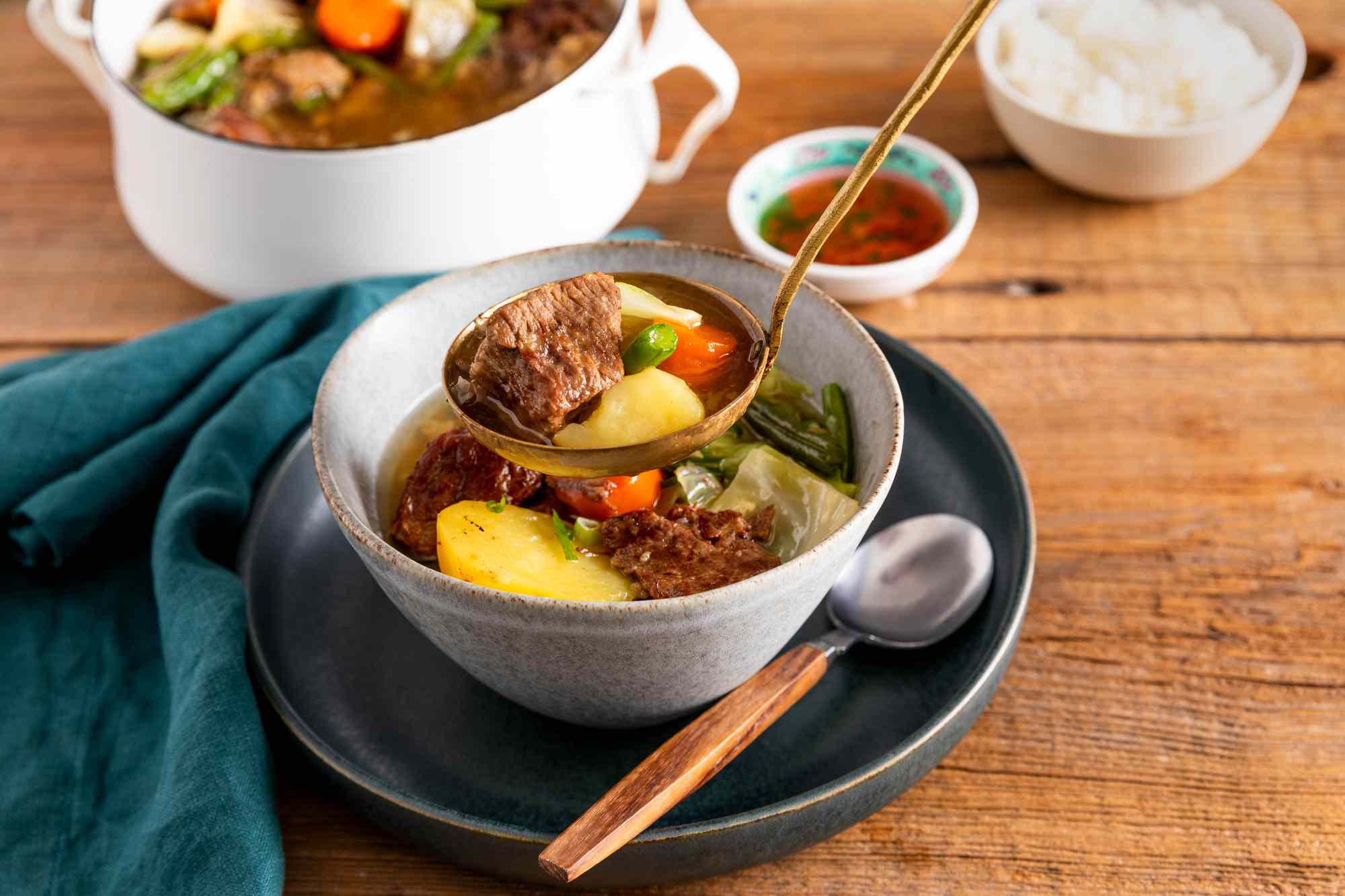Ladle adding Filipino beef stew to a bowl with some stew, and in the surroundings, a bowl of rice, a small bowl of fish sauce, a Dutch oven with more stew, and a navy table napkin