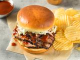 bqq sandwich (slices of juicy meat topped with sauce and coleslaw) next to chips, a bowl of sauce, and a pepper grinder