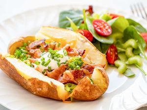 Cheesy, broccoli, bacon loaded baked potato on a white plate with a salad of spinach and tomato behind it on a white plate. The potato is sliced down each side and the top and bottom. Chopped bacon, broccoli, scallions and shredded cheddar cheese are on the potato. A spoonful of sour cream is in the middle of the potato. A fork is to the right of the plate.