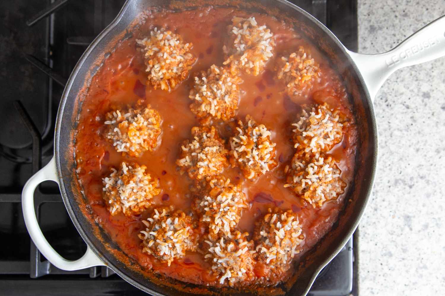 Overhead view of a cast iron skillet on a gas stove after simmering the meatballs and showing the expanded rice on the surface for Easy Porcupine Meatballs recipe