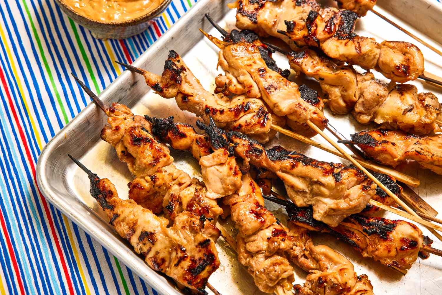 Grilled chicken skewers arranged on a metal tray, sauce bowl in the background