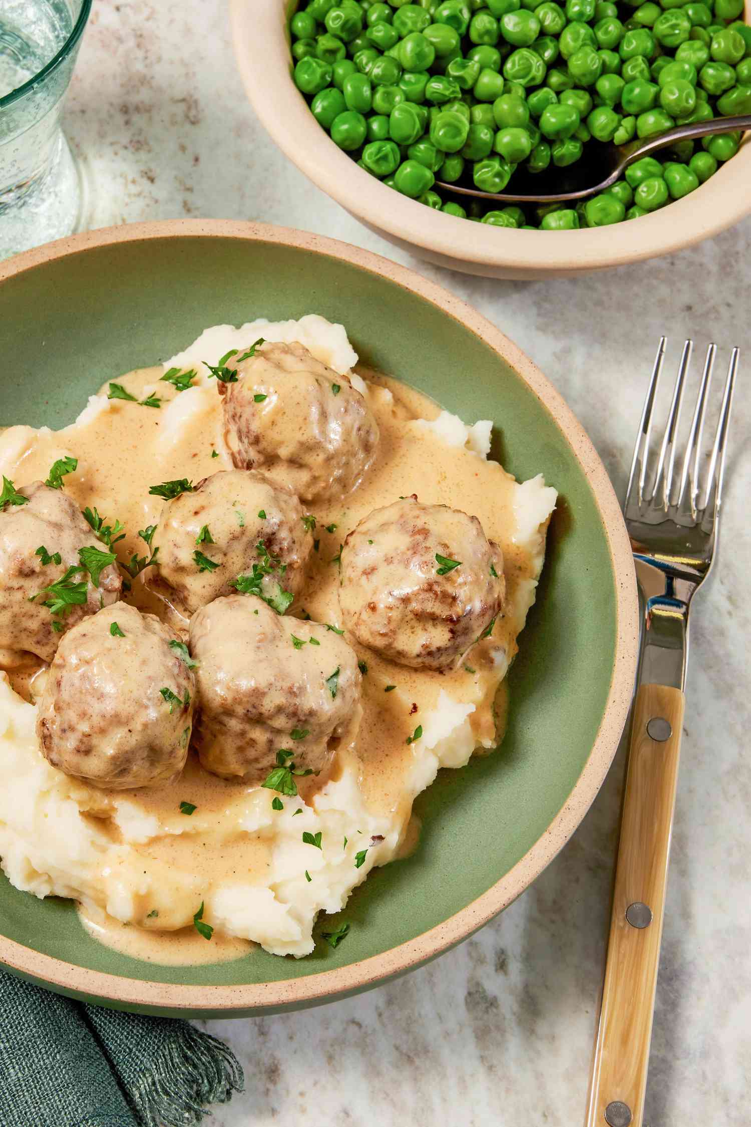 Bowl of copycat Ikea Swedish meatballs served over mashed potatoes, and in the surroundings, a table setting with a bowl of peas, a glass of water, and a sage table napkin