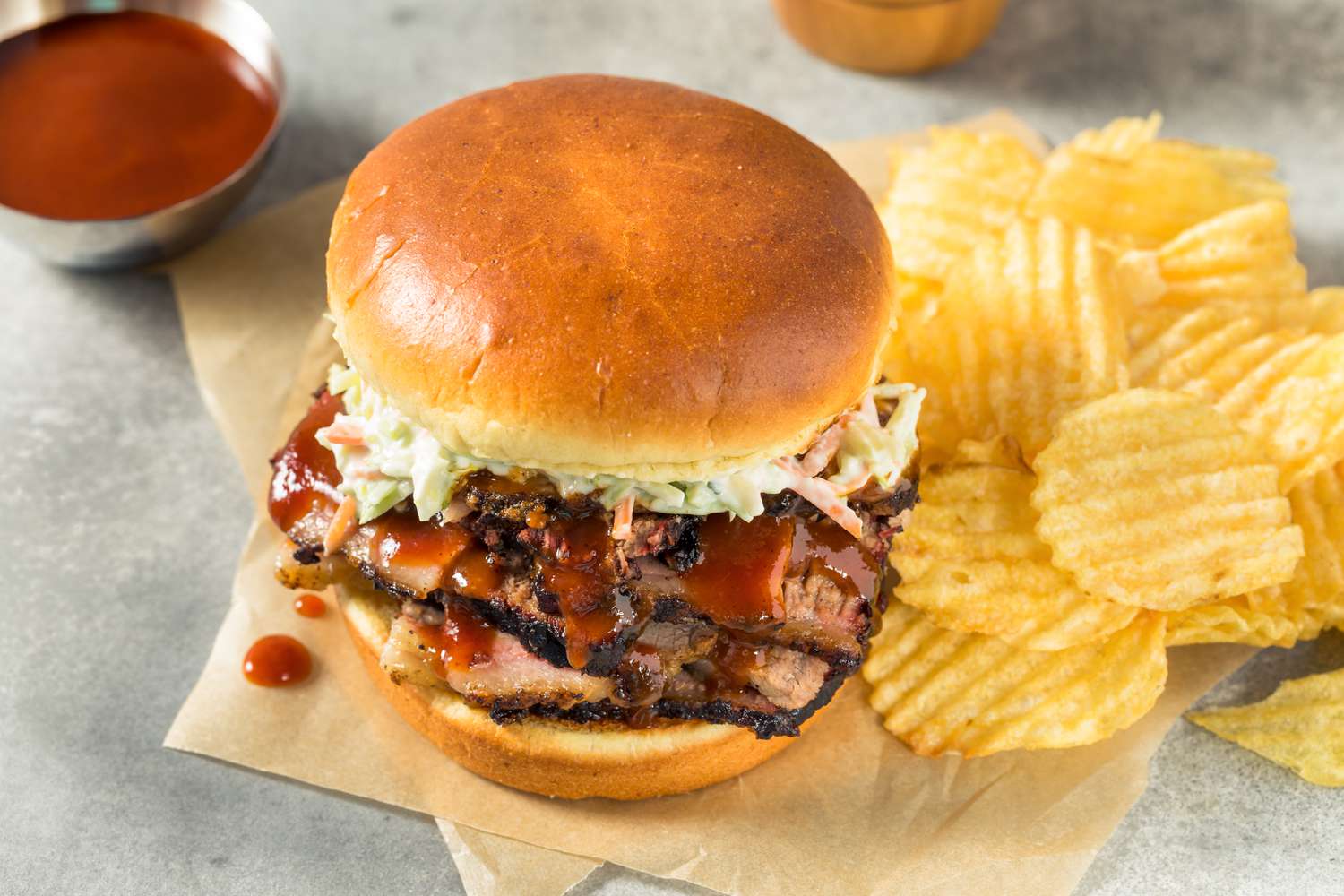 bqq sandwich (slices of juicy meat topped with sauce and coleslaw) next to chips, a bowl of sauce, and a pepper grinder