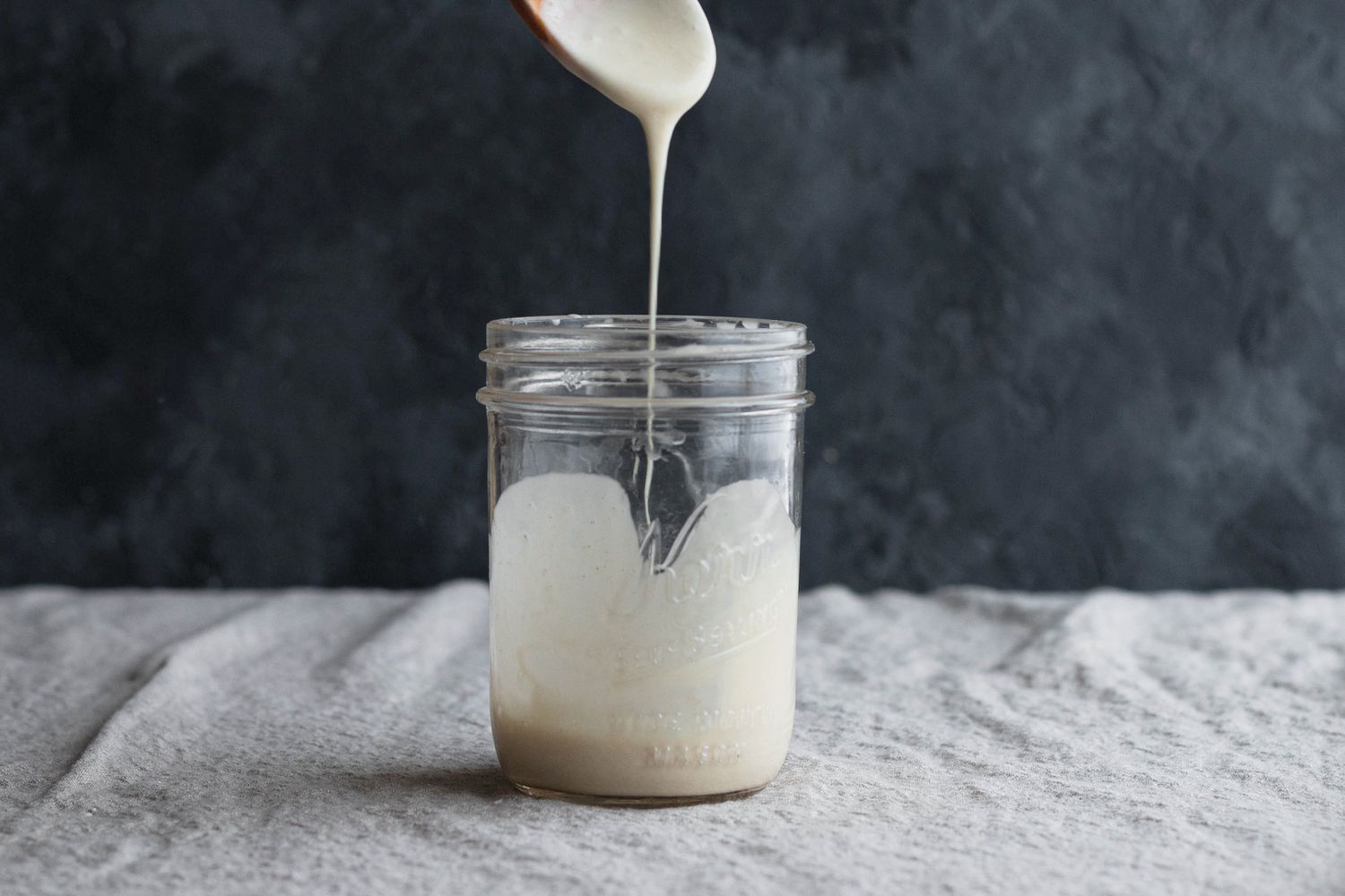 A wooden spoon dripping sourdough starter into a mason jar to show how to make a sourdough starter